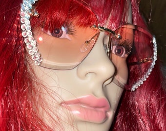 May include: A pair of pink tinted sunglasses with a gold frame and rhinestones on the arms. The sunglasses are on a mannequin with red hair.