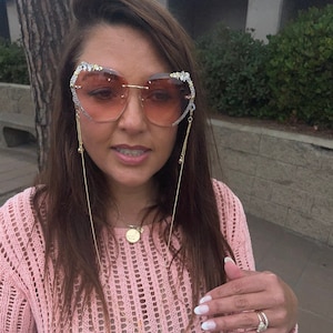 May include: A woman wearing a pink knit sweater with a heart-shaped pair of sunglasses with rhinestones. The sunglasses have a gold chain attached to them. The woman is wearing a gold necklace and a ring on her finger.