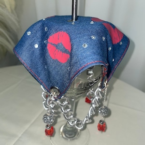 Puede incluir: Un charm para copa de vino de mezclilla azul con labios rojos y detalles de cadena plateada. El charm está unido a un tallo de vidrio con cuentas rojas y plateadas.