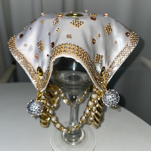 Puede incluir: Una cubierta de copa de vino de satén blanco con adornos dorados y de cristal. La cubierta está drapeada sobre una copa de vino transparente, adornada con cadenas doradas, perlas de imitación y detalles de cristal. El diseño presenta una forma cuadrada con un borde decorativo.