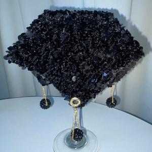 May include: A black sequined fan with gold chains and black bead accents hanging from the bottom. The fan is displayed on a clear glass stand.