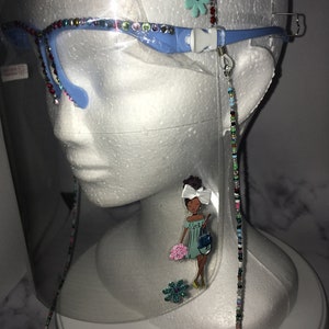 May include: Clear face shield with a blue frame and rhinestones around the edge. A beaded chain is attached to the frame. A cartoon illustration of a woman with a flower and a purse is on the shield.