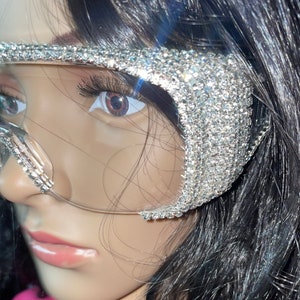 May include: A pair of clear, oversized sunglasses with a silver rhinestone frame. The sunglasses are worn by a person with long black hair.