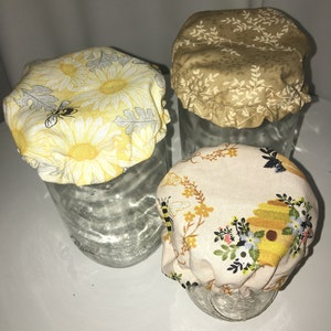 May include: Three glass jars with fabric covers. The covers are made of cotton and feature a bee and flower pattern. The covers are in yellow, brown, and white.