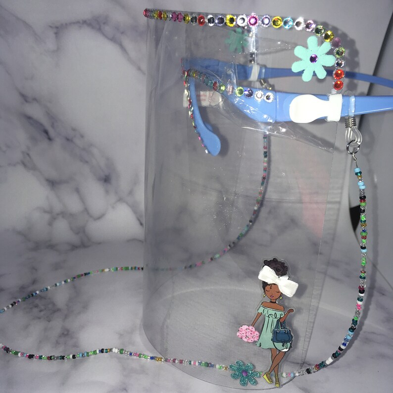 May include: A clear face shield with a blue headband and a beaded chain. The chain has colorful beads and a charm of a cartoon girl with a flower in her hair. The face shield has a rhinestone trim.