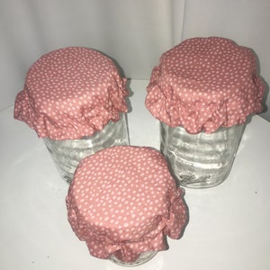 Pode incluir: Três capas de tecido rosa para jarros com bolinhas brancas. As capas têm uma borda franjada e são feitas de um tecido macio e respirável.