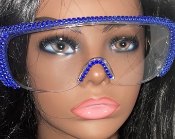 May include: Clear plastic glasses with a blue rhinestone frame. The glasses have a unique shape with a curved top and a straight bottom.