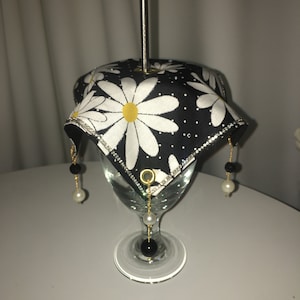 May include: A black and white daisy patterned fabric cover with gold trim and black and white beads hanging from the corners. The cover is placed over a clear glass goblet.