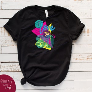 May include: Black t-shirt featuring a graphic design of a colourful geometric pattern and the text "I love the 80's".