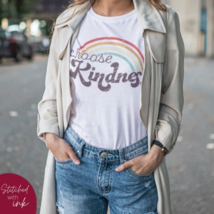May include: White t-shirt with a rainbow graphic and the words "choose Kindness" in a retro font. The shirt is paired with denim shorts and a light-colored trench coat. The image is taken outdoors.