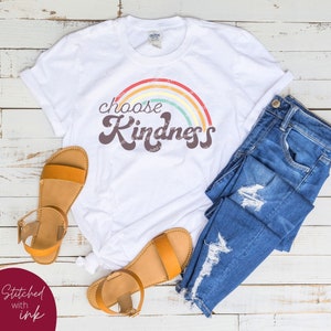 May include: A white t-shirt with the words "choose Kindness" in a retro font, arched over a rainbow design. The shirt is paired with blue denim jeans and brown sandals. The sandals have a strap around the ankle.