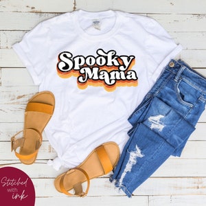 May include: White t-shirt featuring a retro-style graphic that reads "Spooky Mama" in black, orange, and yellow.