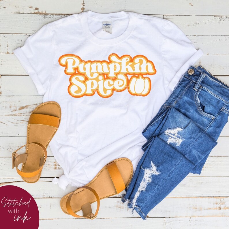 May include: White t-shirt with the text "Pumpkin Spice" in orange and yellow retro font with a pumpkin graphic. The shirt is laid out on a white wooden surface with a pair of brown sandals and blue jeans.