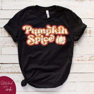 May include: Black t-shirt featuring a retro-style graphic that reads "Pumpkin Spice" in orange and yellow lettering with a brown outline. The text is layered and has a 3D effect.
