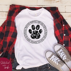 Op de afbeelding: Wit T-shirt met een zwart poot afdruk ontwerp in een cirkel. De cirkel heeft de tekst "DOG MOM" eromheen.