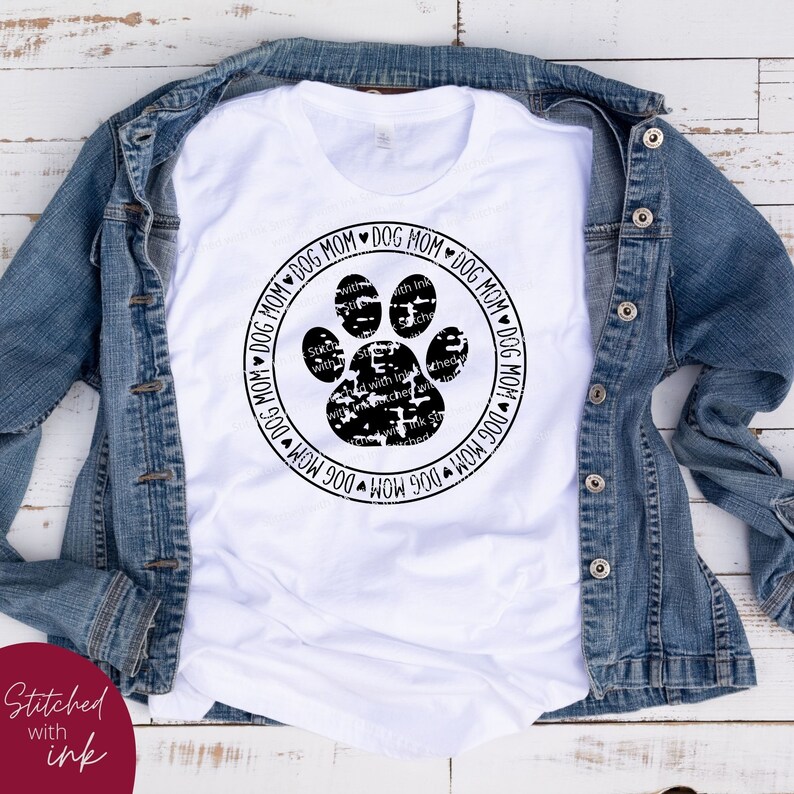 Op de afbeelding: Wit T-shirt met een zwart poot afdruk ontwerp en de tekst "Dog Mom" herhaald rond de poot afdruk.
