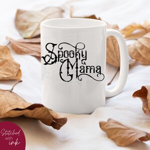 May include: White ceramic mug with a black graphic that says "Spooky Mama" in a stylized font. The mug is sitting on a white surface with brown leaves scattered around it.