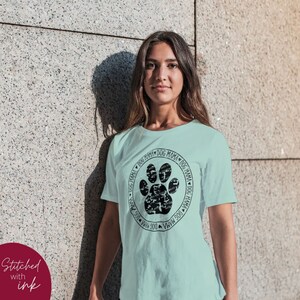 Op de afbeelding: Een lichtblauw T-shirt met een zwart ontwerp van een hondenpoot. De hondenpoot is gemaakt van de woorden "Dog Mama" die in een cirkel worden herhaald.