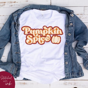May include: White t-shirt featuring a retro-style graphic that reads "Pumpkin Spice" in orange and red lettering with a stylised orange pumpkin.
