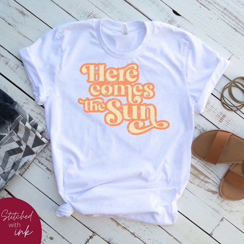 May include: White t-shirt with a peach-colored graphic that says "Here comes the sun" in a retro font.