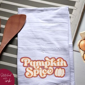 May include: White kitchen towel with a retro-style graphic that says "Pumpkin Spice" in orange and pink letters with a stylized pumpkin.