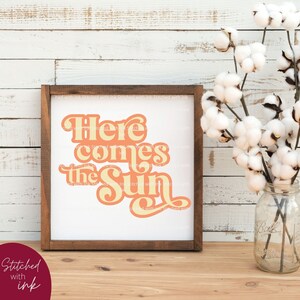 May include: A framed print with a white background and a peach and yellow graphic that says "Here comes the sun". The frame is brown wood and the print is sitting on a wooden surface.