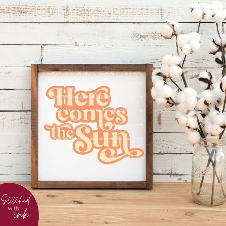 May include: A framed print with a white background and a peach and yellow graphic that says "Here comes the sun". The frame is brown wood and the print is sitting on a wooden surface.
