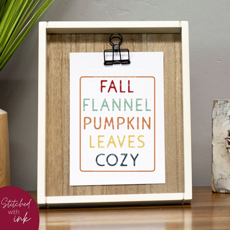 Puede incluir: Una impresi&oacute;n enmarcada con las palabras "Fall, Flannel, Pumpkin, Leaves, Cozy" en un dise&ntilde;o colorido y apilado. La impresi&oacute;n est&aacute; en un marco de madera claro con un passe-partout blanco.