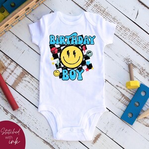May include: White baby onesie with a colorful graphic design featuring a smiley face with lightning bolts, the words "Birthday Boy", and a checkered pattern.
