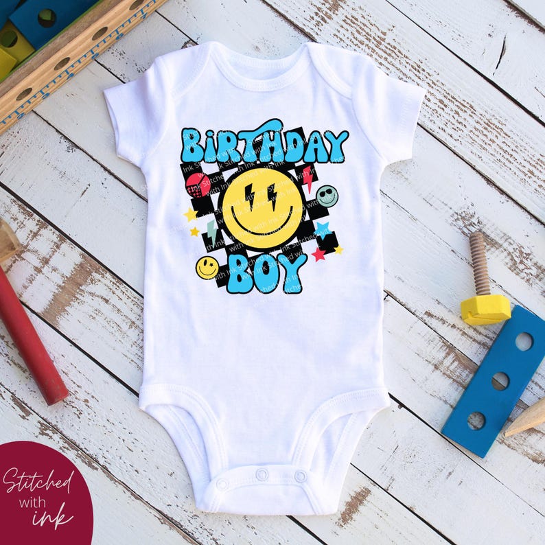 May include: White baby onesie with a colorful graphic design featuring a smiley face with lightning bolts, the words "Birthday Boy", and a checkered pattern.