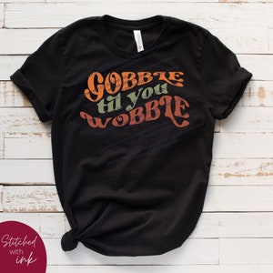 May include: Black t-shirt with a retro-style graphic that says "Gobble til you wobble". The text is in orange, green, and brown.