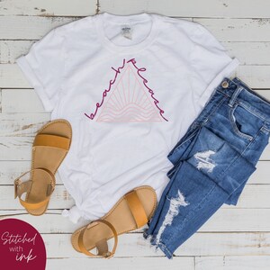 K&ouml;nnte beinhalten: Wei&szlig;es T-Shirt mit einem pinkfarbenen und burgunderroten Grafikdesign eines Dreiecks mit den Worten "beach please" darum herum. Das Shirt liegt auf einer wei&szlig;en Holzoberfl&auml;che mit einem Paar braunen Sandalen und einer blauen Jeans.
