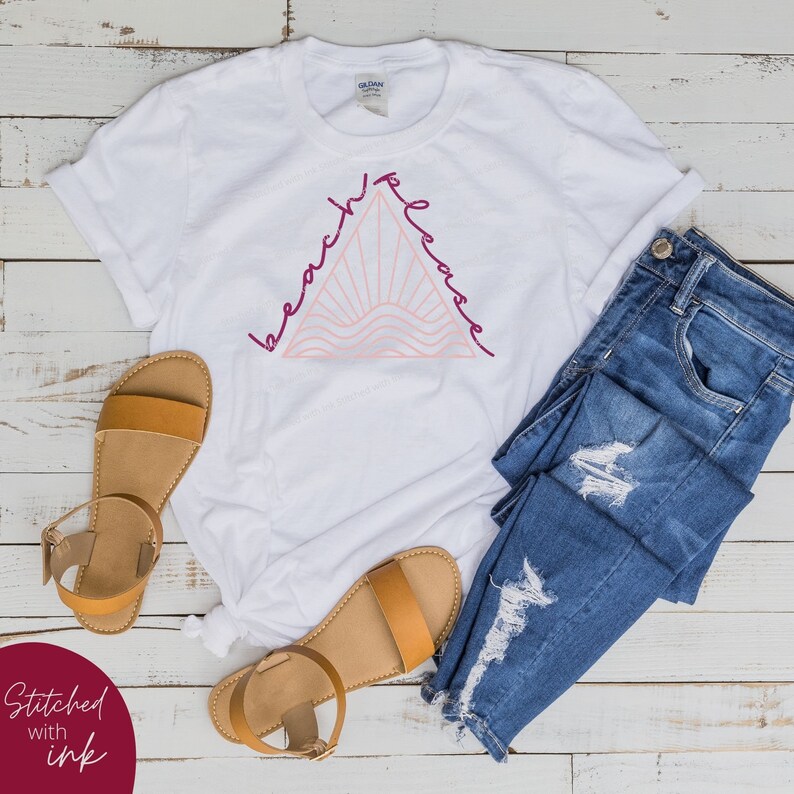 K&ouml;nnte beinhalten: Wei&szlig;es T-Shirt mit einem pinkfarbenen und burgunderroten Grafikdesign eines Dreiecks mit den Worten "beach please" darum herum. Das Shirt liegt auf einer wei&szlig;en Holzoberfl&auml;che mit einem Paar braunen Sandalen und einer blauen Jeans.