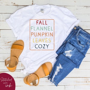 Puede incluir: Camiseta blanca con un gr&aacute;fico de estilo vintage que dice "Fall, Flannel, Pumpkin, Leaves, Cozy". La camiseta est&aacute; colocada sobre una superficie de madera blanca con un par de sandalias marrones y unos vaqueros azules.