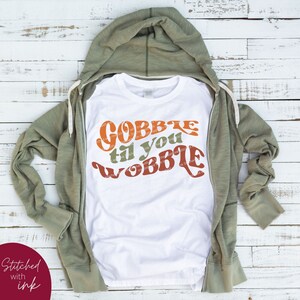 May include: White t-shirt with a retro-style graphic that says "Gobble til you wobble" in orange, brown, and green.