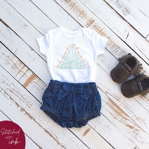 K&ouml;nnte beinhalten: Ein wei&szlig;er Baby-Strampler mit einem gr&uuml;nen Dreiecks-Motiv, auf dem "beach vibes" in einem pfirsichfarbenen Orange steht. Der Strampler wird mit Jeans-Shorts und braunen Mokassins kombiniert.