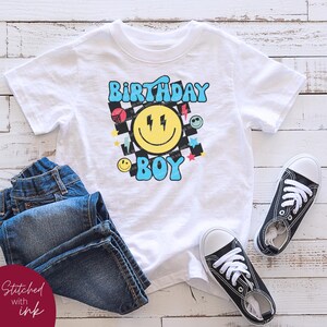 May include: White t-shirt with a blue and yellow smiley face graphic and the text "Birthday Boy" in a retro style. The graphic includes a checkered pattern, lightning bolts, stars, and other colorful shapes.