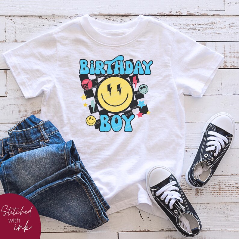 May include: White t-shirt with a blue and yellow smiley face graphic and the text "Birthday Boy" in a retro style. The graphic includes a checkered pattern, lightning bolts, stars, and other colorful shapes.