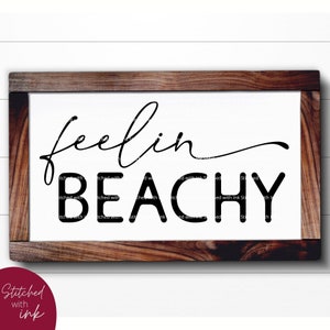 May include: A white wooden sign with a brown frame, featuring the text "feelin beachy" in black cursive font.