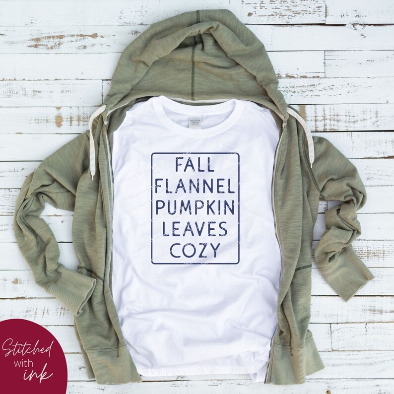 Puede incluir: Camiseta blanca con una caja azul y el texto "FALL FLANNEL PUMPKIN LEAVES COZY" dentro de la caja. La camiseta est&aacute; doblada y colocada sobre una superficie de madera con una sudadera con capucha verde encima.