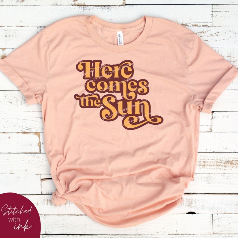 May include: A peach-colored t-shirt with the text "Here comes the sun" in a retro font. The text is in shades of orange and brown.
