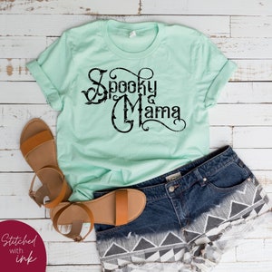 May include: A light green t-shirt with a black graphic that says "Spooky Mama" in a decorative font.