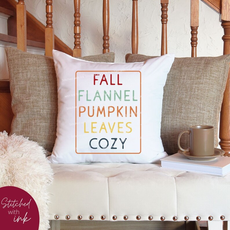 Puede incluir: Coj&iacute;n blanco con un gr&aacute;fico colorido que dice "Fall Flannel Pumpkin Leaves Cozy". El coj&iacute;n est&aacute; en un sof&aacute; blanco con una manta de lana marr&oacute;n y un libro blanco.