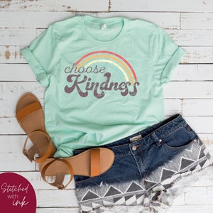 May include: A mint green t-shirt with the words "choose Kindness" in a retro font, above a rainbow design. The shirt is paired with denim shorts and brown sandals. The words "Stitched with ink" are in the lower left corner.