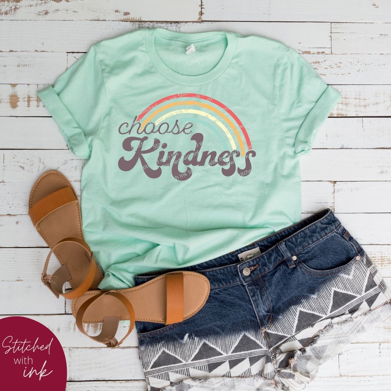 May include: A mint green t-shirt with the words "choose Kindness" in a retro font, above a rainbow design. The shirt is paired with denim shorts and brown sandals. The words "Stitched with ink" are in the lower left corner.