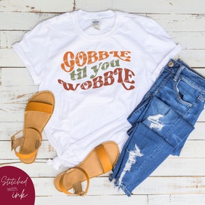 May include: White t-shirt with the text "Gobble til you wobble" in a retro font with orange, green, and brown colors. The shirt is displayed on a wooden surface with a pair of brown sandals and blue jeans.