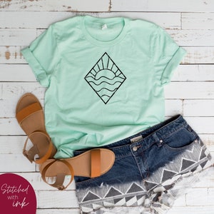 May include: A light green t-shirt with a black graphic of a sun setting over waves inside a diamond shape. The t-shirt is paired with denim shorts and brown sandals.