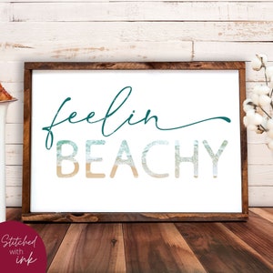 May include: A framed sign with the text "feelin BEACHY" in teal and white lettering. The letters are filled with a beach scene of sand and ocean waves.