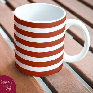 K&ouml;nnte beinhalten: Eine wei&szlig;e Keramiktasse mit einem rot-wei&szlig; gestreiften Design. Die Tasse hat einen Henkel und steht auf einer Holzoberfl&auml;che.