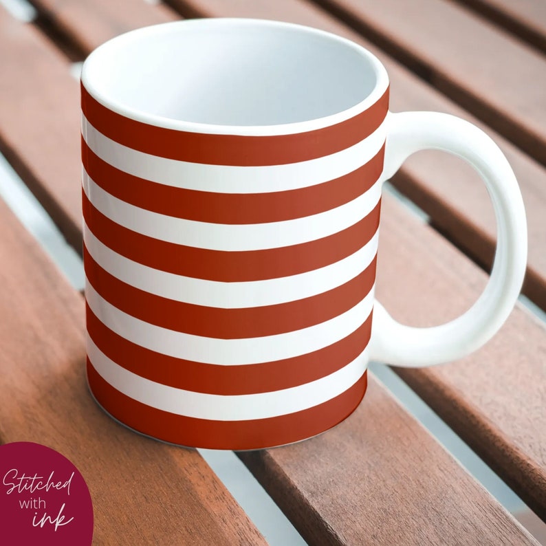 K&ouml;nnte beinhalten: Eine wei&szlig;e Keramiktasse mit einem rot-wei&szlig; gestreiften Design. Die Tasse hat einen Henkel und steht auf einer Holzoberfl&auml;che.